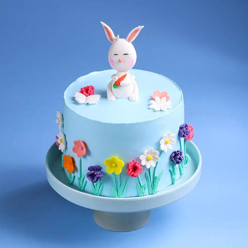 Light blue birthday cake with a 3D fondant bunny topper holding a carrot and colorful gumpaste flowers on the sides.