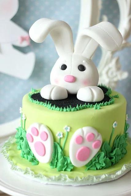 A light green fondant cake featuring a 3D white bunny topper emerging from a black hole with pink paw prints and green icing grass.