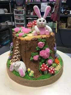 A brown tree stump cake with a white fondant bunny on top, pink roses, and red mushrooms on a green grass base.