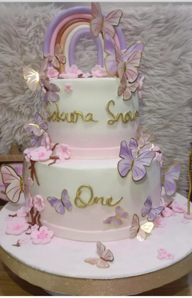 Two-tier white and pink ombre fondant cake decorated with purple butterflies, pink cherry blossoms, and a pastel rainbow topper.