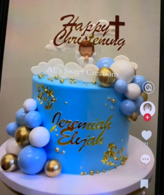 Light blue christening cake with a praying angel figurine, gold and blue plastic balls, and white fondant clouds.