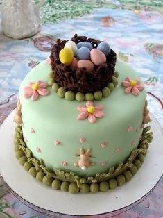 A light green fondant cake featuring a chocolate bird nest with colorful eggs on top, pink flowers, and a small bunny at the base.