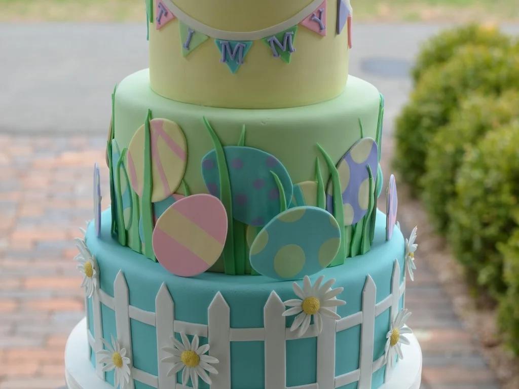 Three-tier pastel Easter themed cake with yellow, green, and blue fondant tiers featuring a white picket fence, daisies, and colorful eggs.