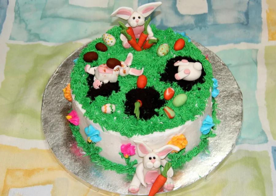 White round cake with green grass icing, fondant bunnies, carrots, and colorful Easter eggs on a silver base.