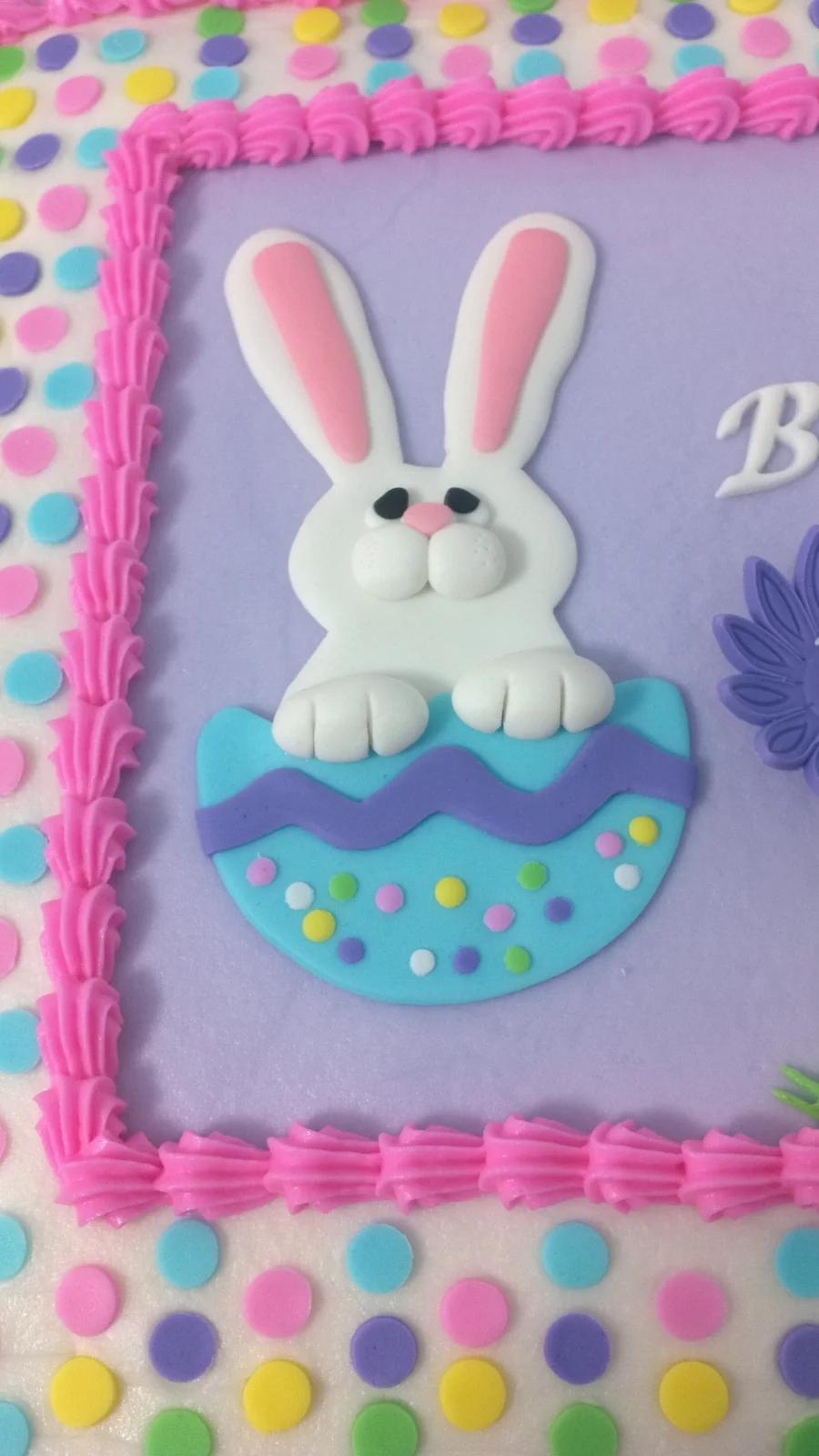 Square cake with a purple icing center, pink piped borders, and a 3D fondant bunny in a blue decorated Easter egg.