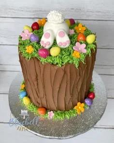 A tall chocolate brown cake textured like a tree stump with green icing grass, colorful fondant eggs, and a white bunny butt topper.