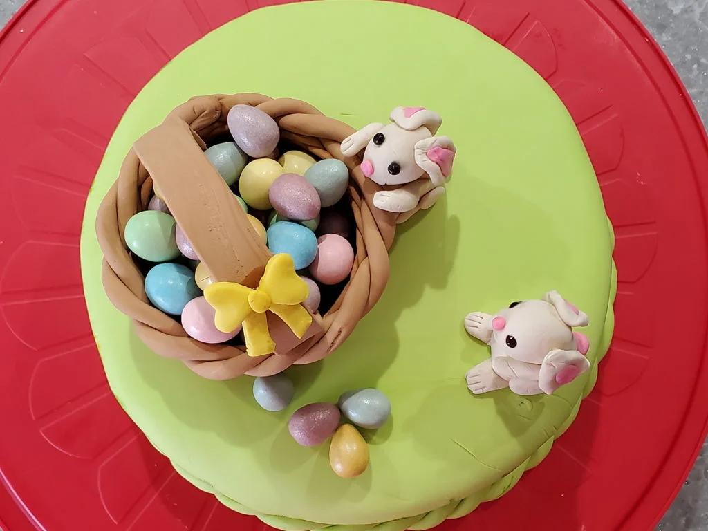 Easter themed green fondant cake featuring a brown woven basket filled with colorful pastel eggs and two white fondant bunnies.