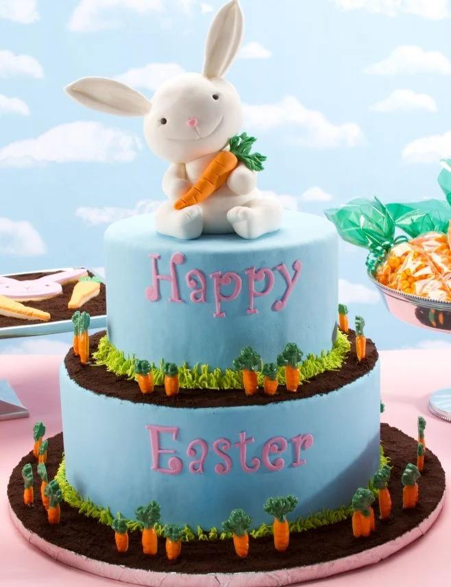 Two-tier light blue fondant Easter cake featuring a large white bunny topper holding a carrot, surrounded by small fondant carrots and grass.