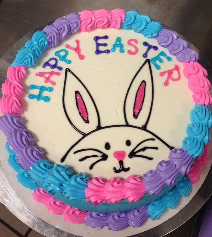 White round cake with colorful piped icing borders and a hand-drawn icing bunny face with Happy Easter message.