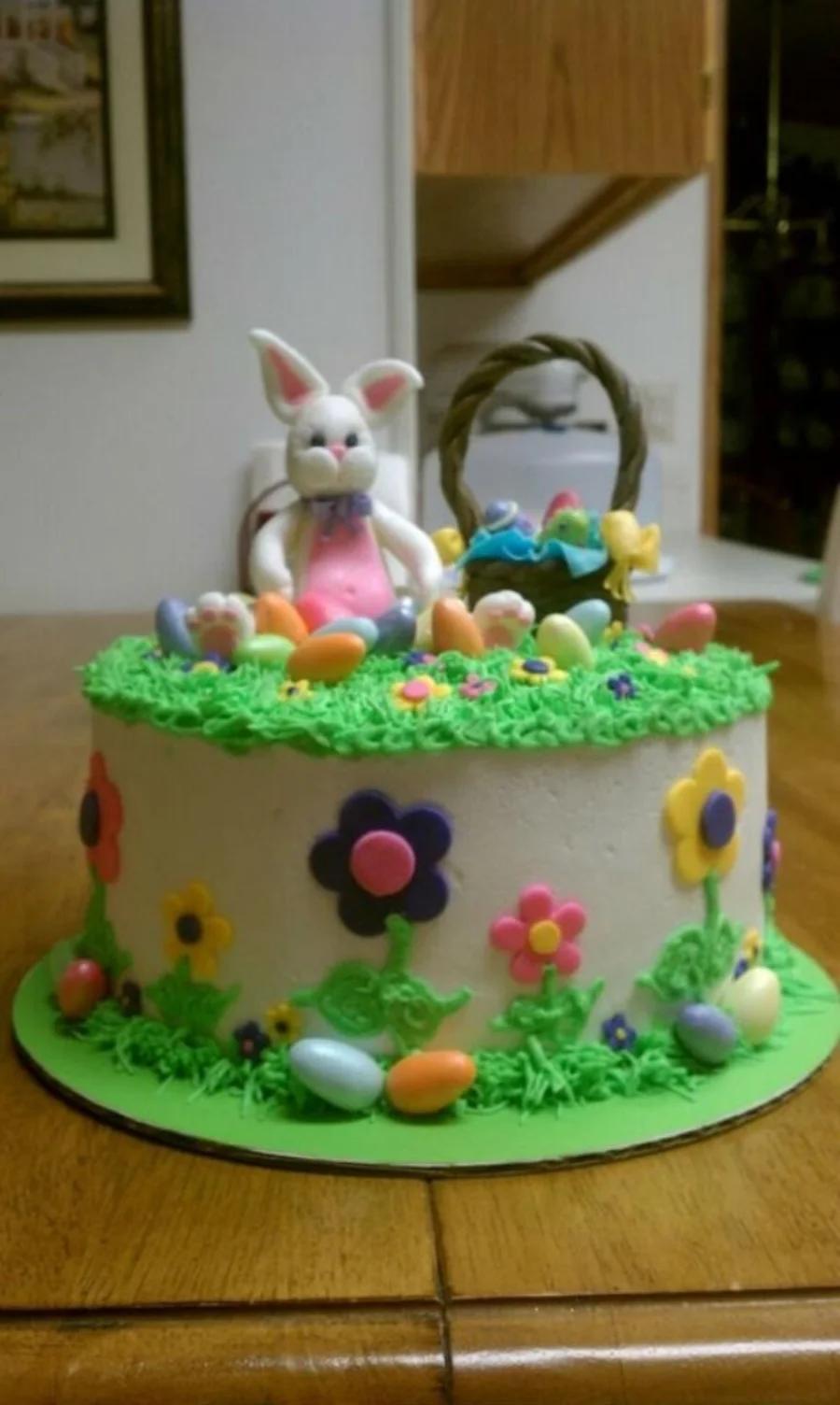White 1-tier Easter cake with a 3D fondant bunny, a basket of eggs, and colorful icing flowers and grass.