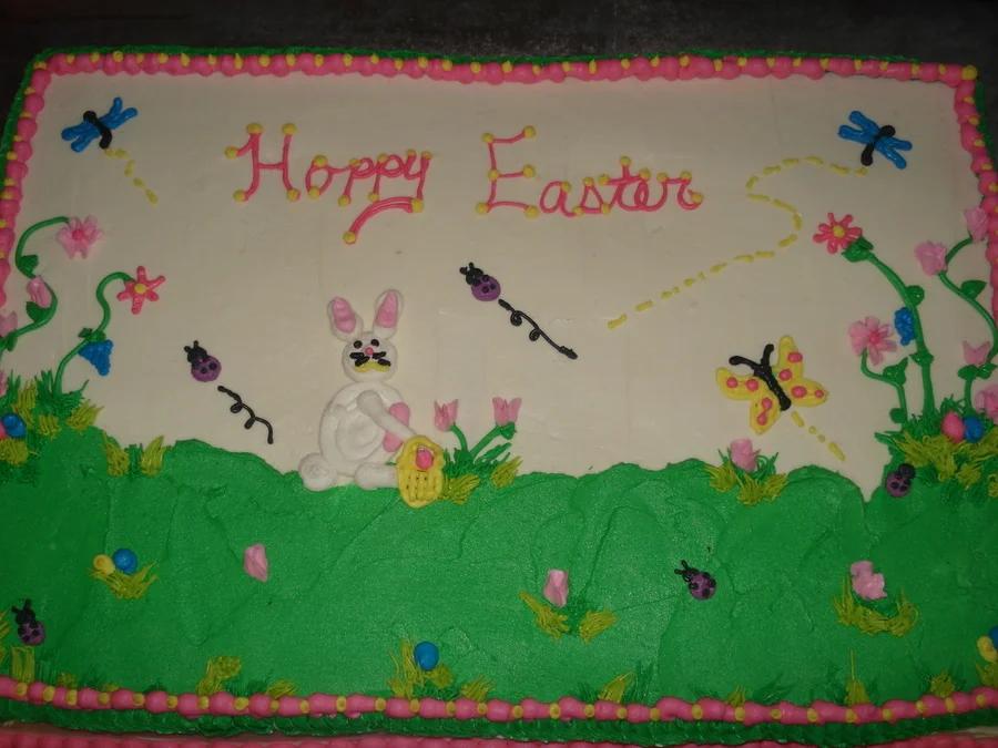 Rectangular Easter sheet cake with white icing, green grass, a white fondant bunny, and colorful icing flowers and insects.
