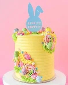 Tall yellow ribbed buttercream cake with colorful icing rosettes, speckled Easter eggs, and a blue bunny ear topper.