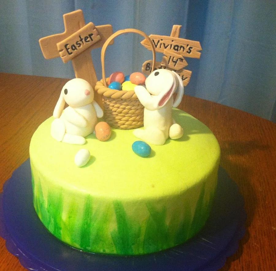 Easter themed birthday cake with fondant bunnies, a basket of colorful eggs, and wooden-style signs on a green grass-painted base.