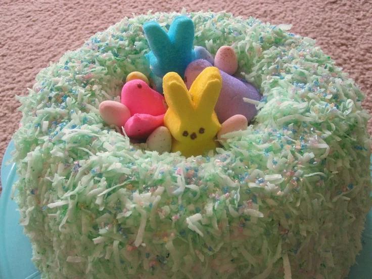 Easter nest cake covered in light green coconut flakes with colorful marshmallow Peeps bunnies and speckled candy eggs on top.