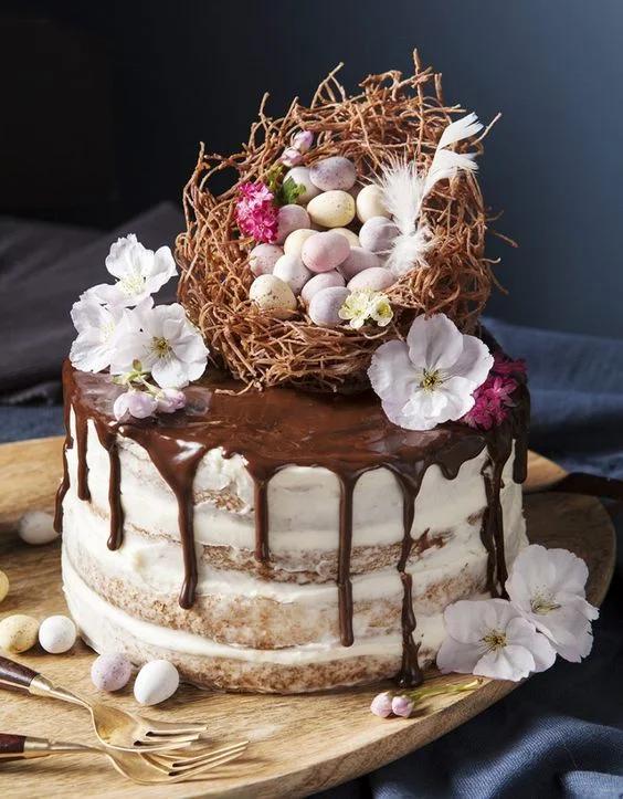 Naked cake with chocolate drip, topped with a bird's nest made of shredded pastry, speckled eggs, and white cherry blossoms.
