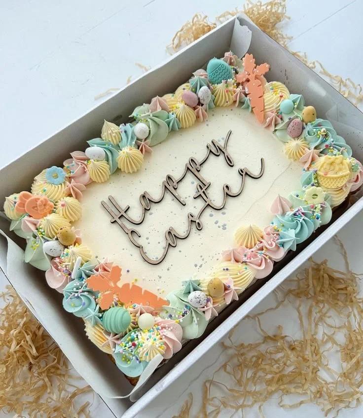 Rectangular Easter cake with white soft icing, pastel buttercream rosettes, chocolate eggs, and a wooden Happy Easter topper.