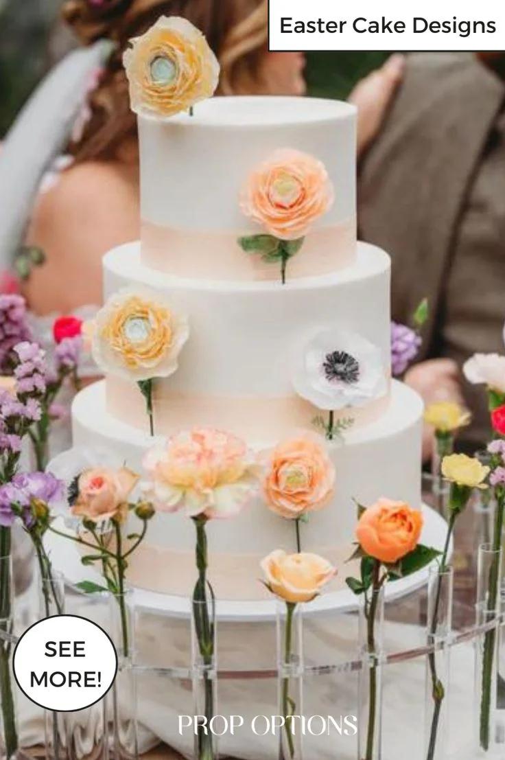 Elegant 3-tier white fondant wedding cake decorated with peach, ivory, and white ranunculus and anemone flowers on a clear stand.