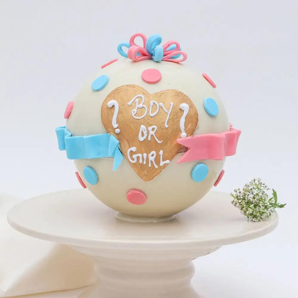 Spherical white gender reveal cake with pink and blue fondant dots, a gold heart, and 'Boy or Girl' message.