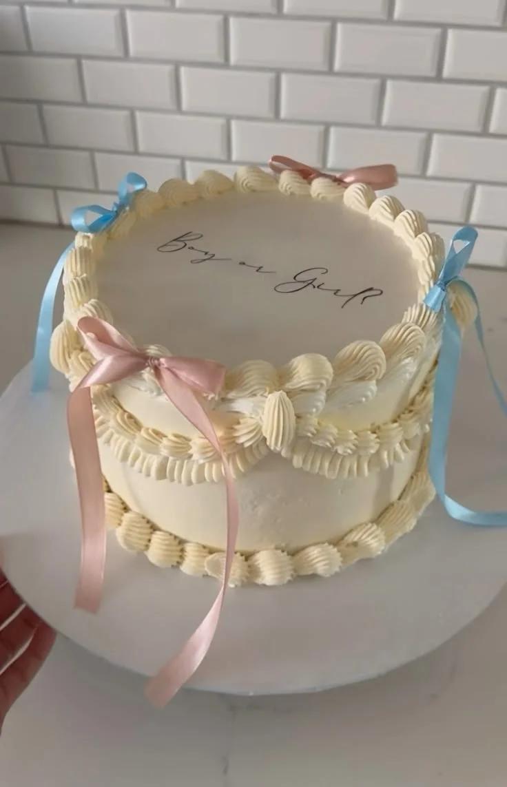 Elegant cream-colored vintage-style gender reveal cake with pink and blue satin ribbons and 'Boy or Girl' icing text.