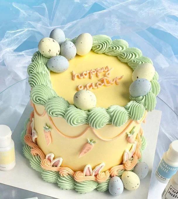 Yellow Easter cake with light green ruffled icing borders, fondant carrots, bunny ears, and speckled pastel chocolate eggs.