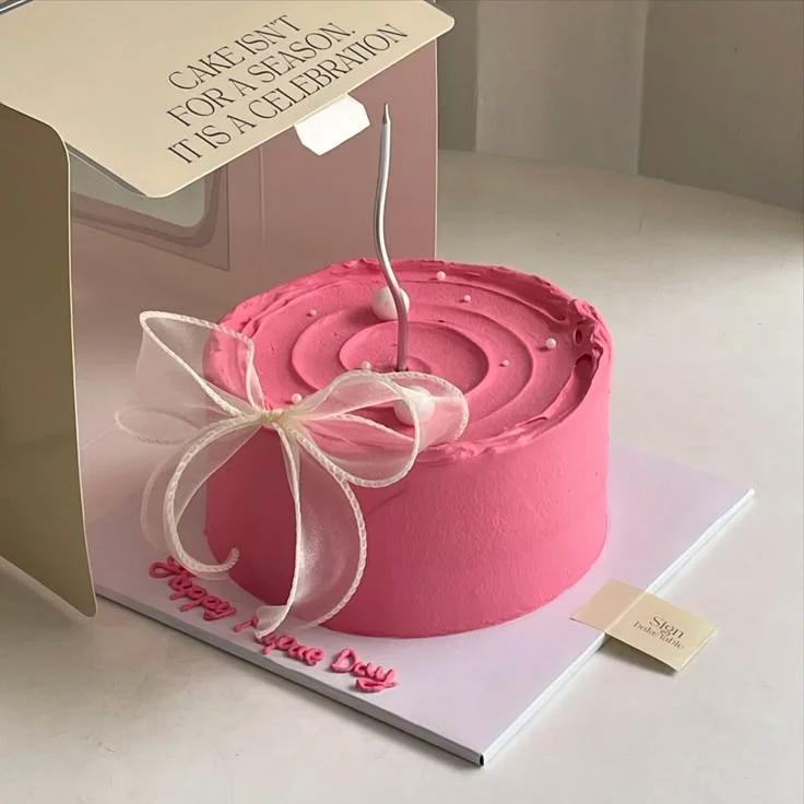 Minimalist hot pink cake with a tall silver candle, white pearls, and a large white sheer ribbon bow on a white baseboard.