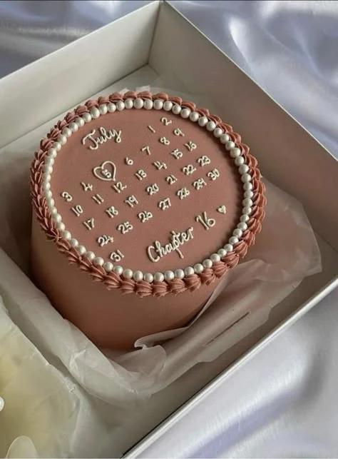 A minimalist dusty rose pink cake featuring a white piped icing calendar for July with 'Chapter 16' and a heart around the 5th.
