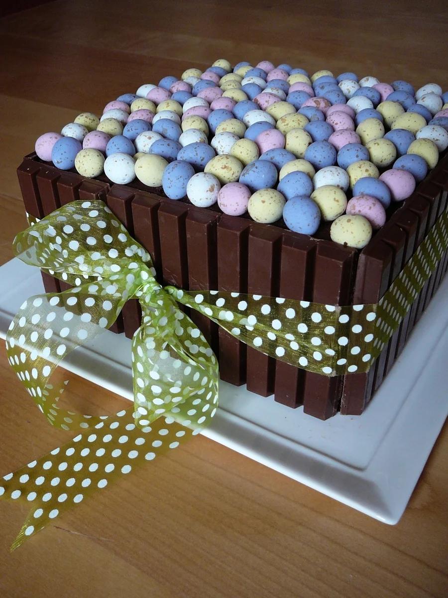 Square chocolate cake surrounded by KitKat bars and topped with colorful pastel candy eggs, finished with a green polka dot ribbon.