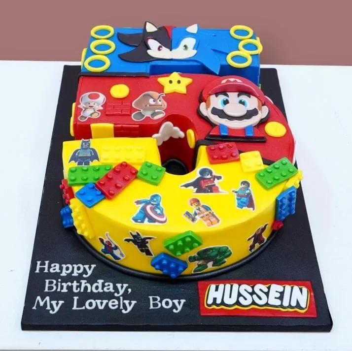 Number 5 shaped fondant cake featuring Super Mario, Sonic the Hedgehog, and Lego superhero characters with colorful Lego brick decorations.