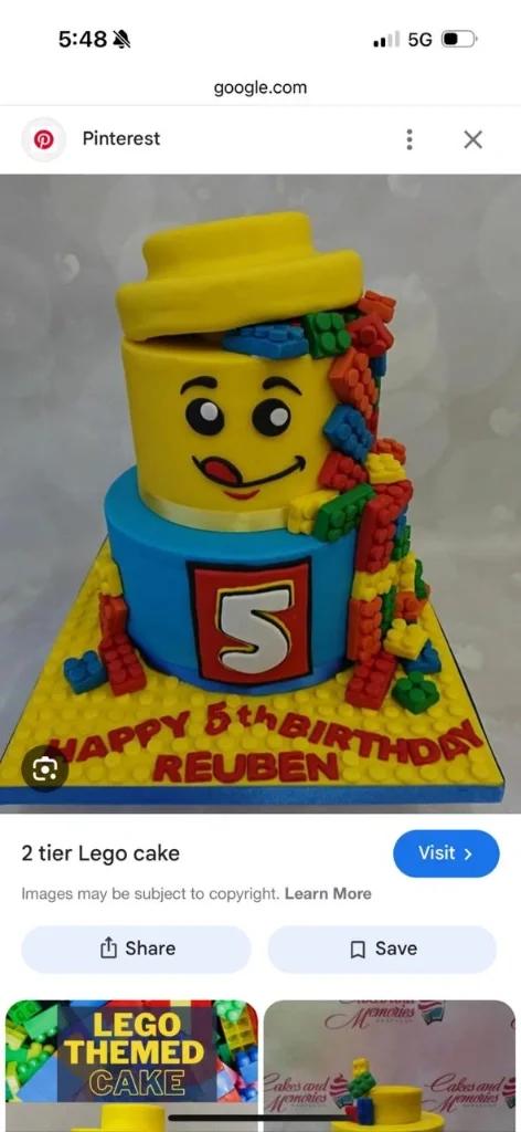 Two-tier Lego themed birthday cake featuring a yellow Lego head top tier and blue bottom tier with colorful building blocks cascading down.