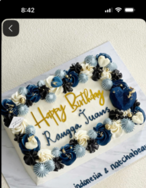 A rectangular white soft-icing cake with navy blue, black, and light blue icing rosettes, gold dragees, and a gold Happy Birthday topper.