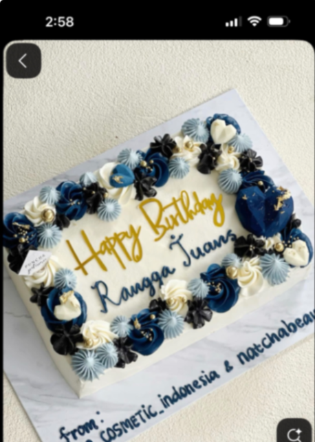 A rectangular white soft-icing cake with a border of navy blue, light blue, and white icing rosettes, gold dragees, and a blue geometric heart.