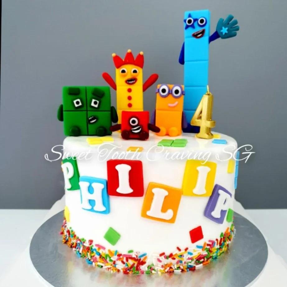 White fondant Numberblocks themed birthday cake with colorful 3D character toppers, rainbow sprinkles, and a gold number 4 candle.