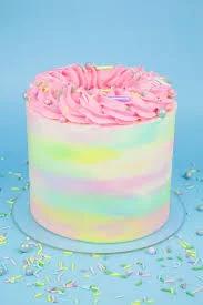 A tall single-tier cake with pastel rainbow watercolor icing, topped with a thick pink icing swirl and colorful sprinkles.