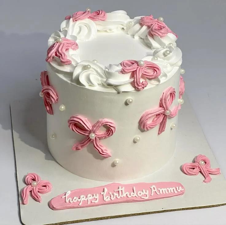 White minimalist cake with pink piped icing bows and white pearls, featuring a 'Happy Birthday Ammu' message on a pink icing strip.