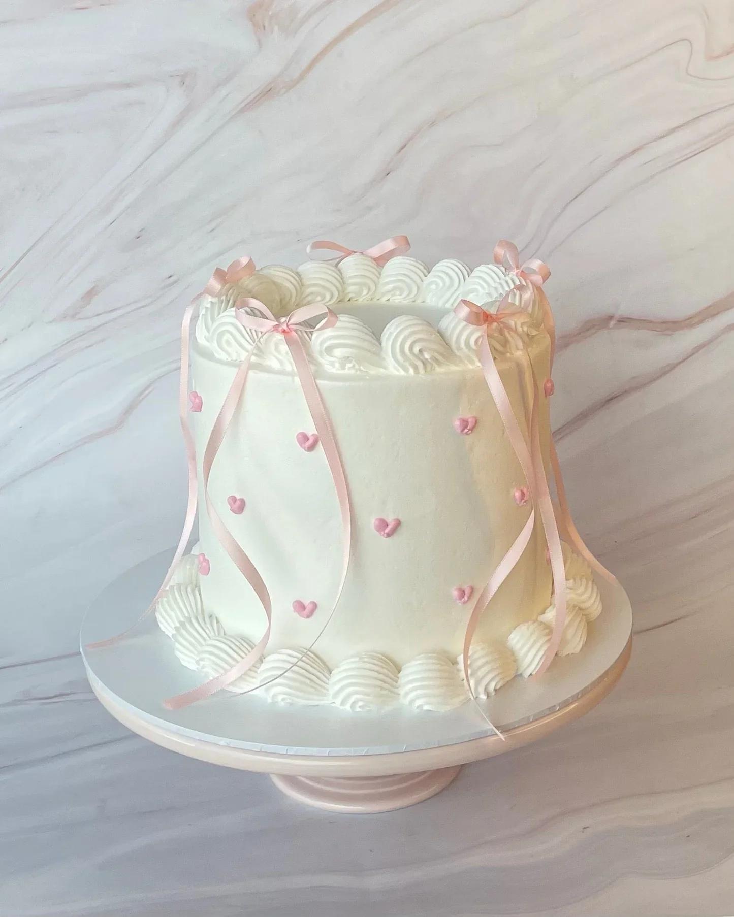 Tall white minimalist cake with pink piped icing hearts and delicate pink satin ribbons on a white cake stand.