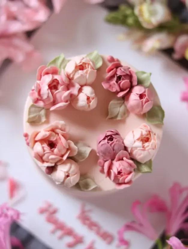 A round light pink cake decorated with realistic pink and peach edible gumpaste peonies and green leaves on top.