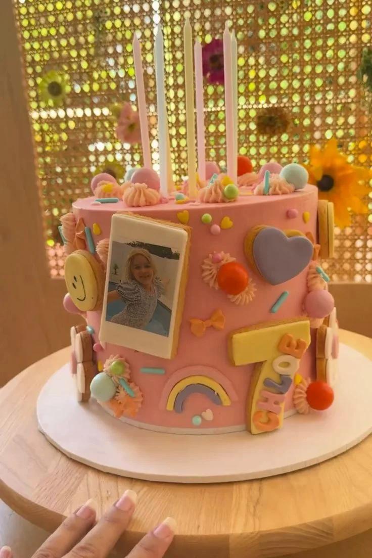 Pink birthday cake with a polaroid photo printout, rainbow, heart, and smiley face cookie toppers with tall pastel candles.