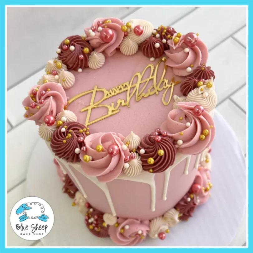 Pink 1-tier birthday cake with pink and maroon icing rosettes, white chocolate drip, and a gold Happy Birthday topper.