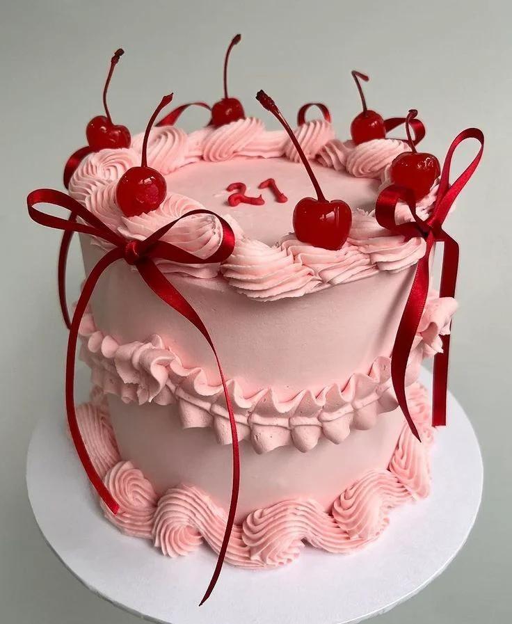 Pink vintage-style heart-shaped cake with piped ruffles, red cherries, and red satin bows for a 21st birthday.