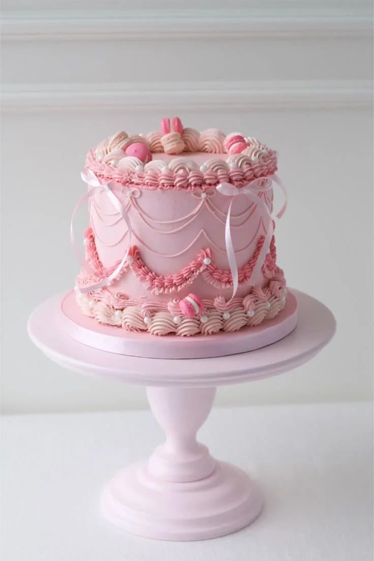 Elegant pink vintage-style cake with intricate ruffles, piped swags, white pearls, and pink macarons on a pink cake stand.