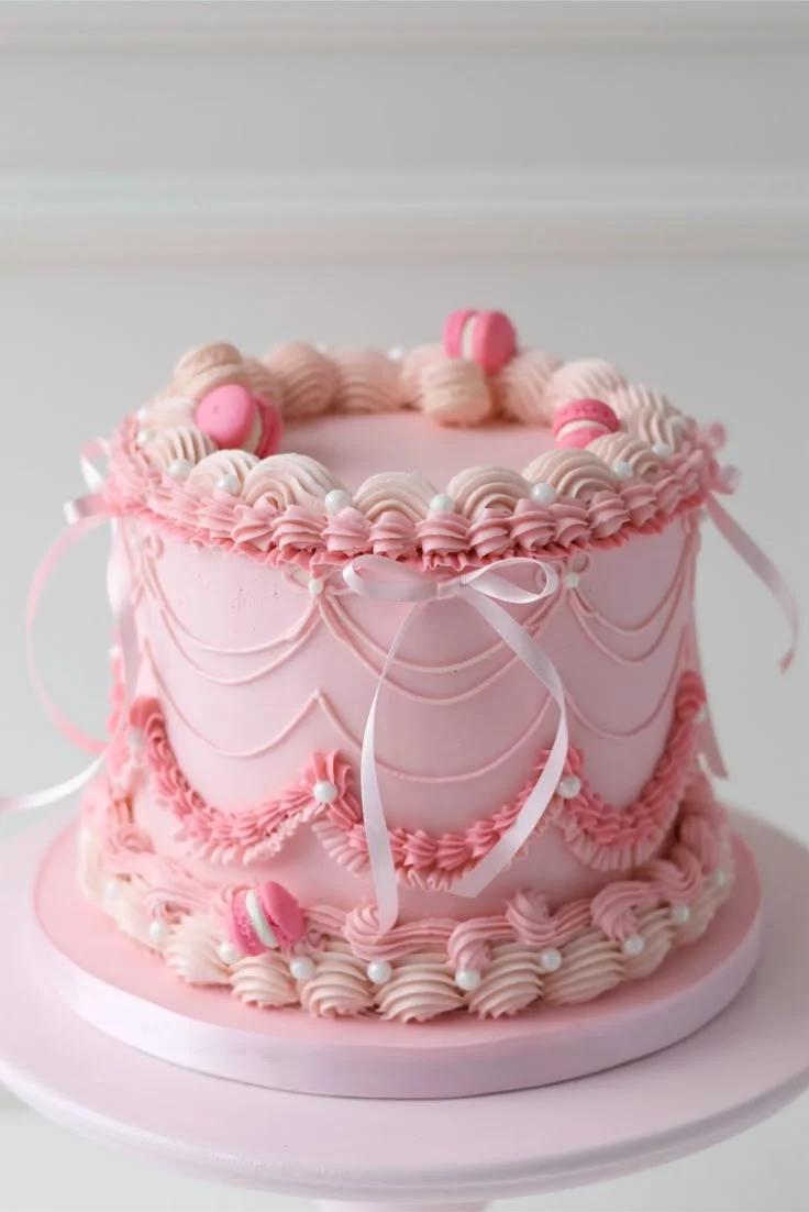 Elegant pink vintage-style cake with intricate piped ruffles, swags, and pink macarons on a matching pink base.