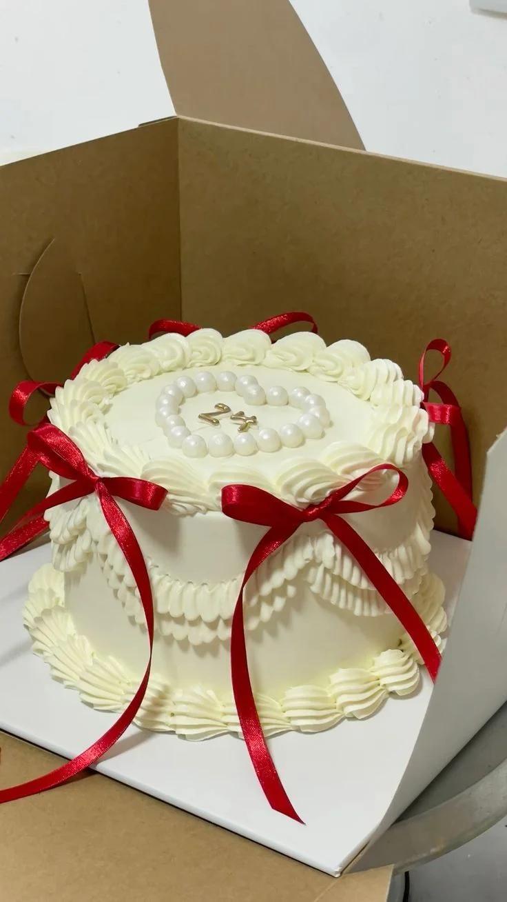 White minimalist vintage cake with red satin bows, piped cream ruffles, and a pearl heart with gold number 24 on top.