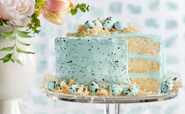 Light blue robin egg inspired cake with chocolate speckles, topped with blue eggs and edible nest on a glass stand.