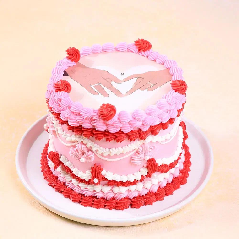 Pink vintage heart-themed cake with intricate pink, red, and white icing ruffles and a romantic hand-heart printout topper.