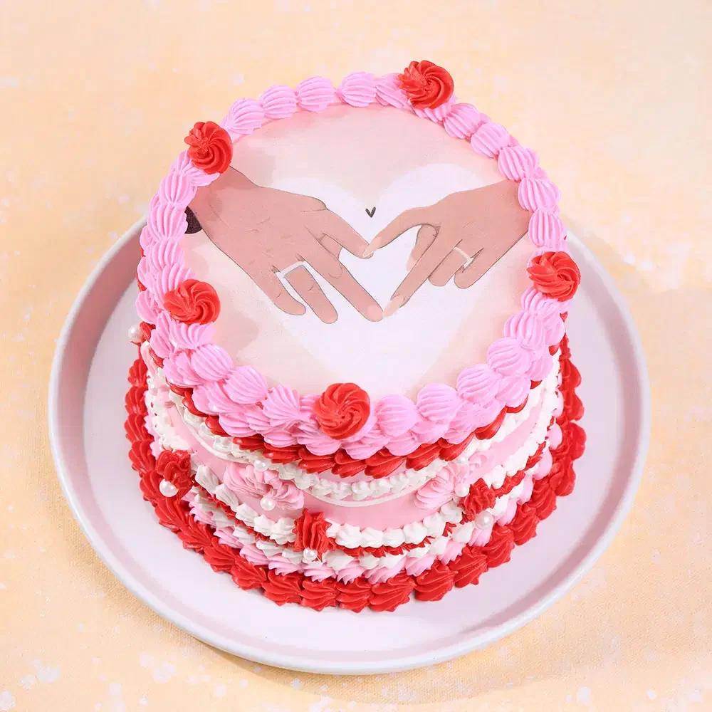 Pink and red vintage-style heart cake with a printed topper of two hands forming a heart shape and intricate icing ruffles.