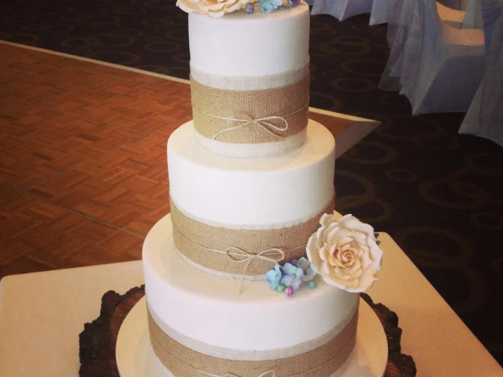 Elegant 3-tier white fondant wedding cake with rustic burlap ribbons, twine bows, and delicate peach and blue sugar flowers.