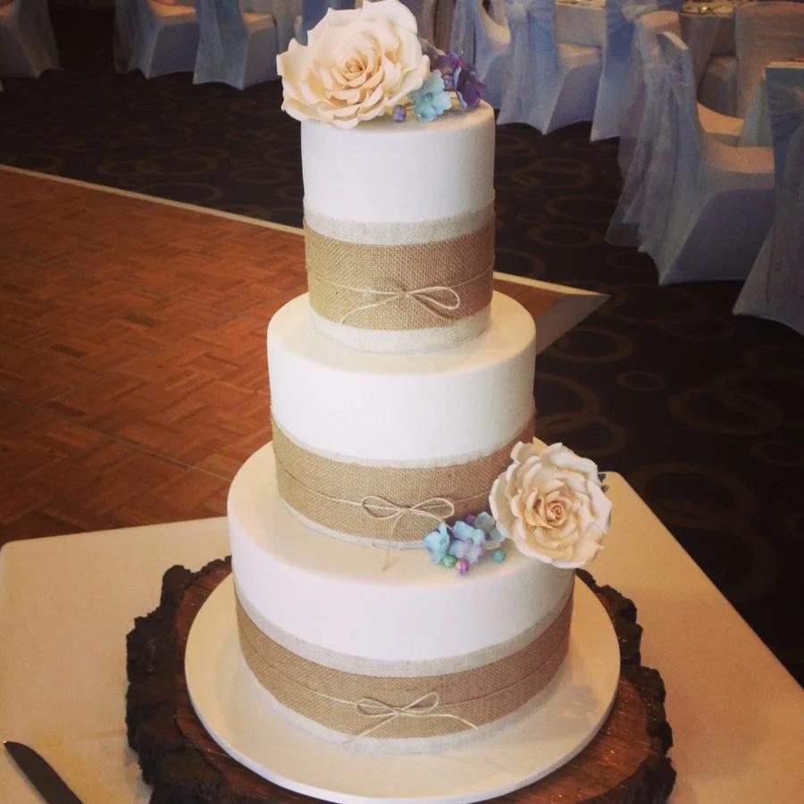 Elegant three-tier white fondant wedding cake featuring burlap ribbons, twine bows, and large ivory and blue gumpaste roses on a wood slab.