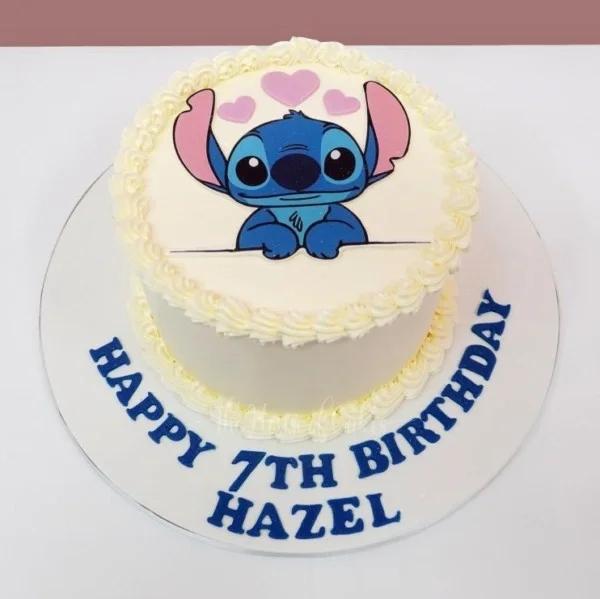 White round cake with a blue Stitch character printout on top, pink hearts, and blue icing text on the base board.