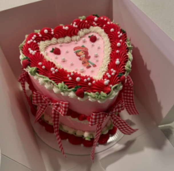 Heart-shaped pink Strawberry Shortcake themed cake with red and white icing ruffles, gingham bows, and a character printout. Heart-shaped pink Strawberry Shortcake themed cake with red and white icing ruffles, gingham bows, and a character printout.