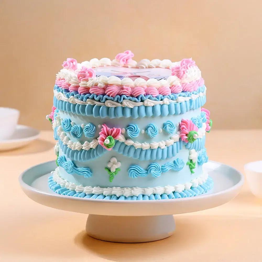 Light blue vintage style cake with intricate pink and white icing ruffles, rosettes, and a printed character topper on top.